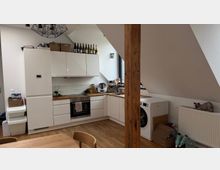 Helle Küchenzeile in einem Dachgeschoss mit weißen Schränken, Holzarbeitsplatten und einer schrägen Decke. Links ein Kühlschrank, in der Mitte ein Herd mit Backofen und rechts eine Waschmaschine; ein freistehender Holzbalken steht zentral im Raum.