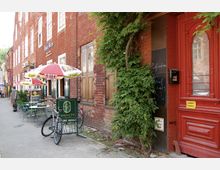 Straßenansicht einer roten Backsteinfassade mit großen Fenstern und einer roten Holztür. Im Vordergrund steht ein Café mit Außensitzplätzen, Sonnenschirmen und einem Fahrradständer; Pflanzen ranken an der Wand empor.