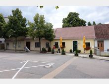 Straßenszene mit einstöckigen Häusern mit roten Ziegeldächern und Fenstern mit Blumenkästen; vor einem Haus stehen zwei gelbe Sonnenschirme. Im Vordergrund Gehwege und eine Straße, zwei Menschen gehen und eine Person im Rollstuhl wird begleitet, umgeben von Bäumen.