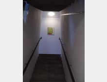 Betontreppe in einem schmalen, minimalistisch gestalteten Treppenhaus mit weißen Wänden und Metallgeländern; an der oberen Wand hängt ein kleines modernes Kunstwerk unter einer Deckenleuchte.