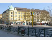 Straßenszene mit einem großen, gelben Gebäude mit Kuppeldach und Geschäften im Erdgeschoss, darunter eine Filiale der Deutschen Bank. Im Vordergrund sind Fahrräder an einem Metallgeländer befestigt, eine Straße verläuft quer durch das Bild, und ein U-Bahn-Schild der Linie U9 ragt in die Höhe.