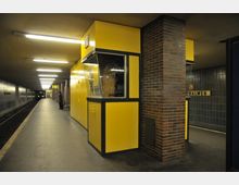 Eine U-Bahn-Station mit gelben Wandverkleidungen und einem Informationsschalter, flankiert von einer Backsteinsäule. Die Bahnsteigkante verläuft entlang der Gleise, und der Bahnhof ist mit kargen Beleuchtungen und grauen Fliesen gestaltet.