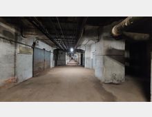 Langer, unterirdischer Gang mit grauen Betonwänden und freiliegenden Rohren und Kabeln an der Decke. Die Umgebung wirkt industriell, mit großen Metalltüren an einer Seite und schwacher Beleuchtung durch wenige Deckenlampen.