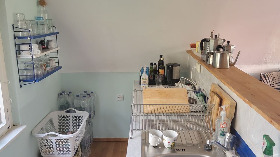 Kleine Küchenzeile in einem schmalen Raum mit Holzfußboden, weiß gestrichener Decke mit sichtbaren Balken und einer Hängelampe. Rechts sind Spüle, Abtropfgestell und Arbeitsfläche mit Küchenutensilien, links ein offenes Wandregal mit Gläsern sowie Wasserkisten am Boden unter einem Fenster.