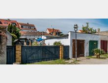 Wohngebiet mit einem Garten und Garagen in ländlicher Umgebung. Im Vordergrund ein Zaun aus dunklem Holz, daneben eine weiße Mauer mit Garagentüren in verschiedenen Farben; im Hintergrund mehrstöckige Häuser mit roten Ziegeldächern.