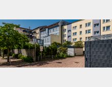 Wohngebäude mit mehreren Etagen in einer ruhigen Wohngegend, umgeben von Bäumen, Sträuchern und einem gepflasterten Weg. Im Vordergrund sind ein Metallzaun, ein Schild mit Parkplatzhinweis und Abstellflächen für Fahrräder zu sehen.