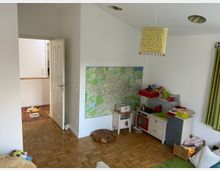 Ein kindgerecht eingerichtetes Zimmer mit Holzboden, einer kleinen Spielküche auf der rechten Seite und einer großen Landkarte an der weißen Wand dahinter. Im Vordergrund sind ein Teppich und Spielzeug zu sehen, während eine offene Tür in einen angrenzenden Raum führt.