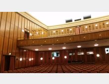 Innenraum eines Konzertsaals mit hölzernen Wandverkleidungen, runden Wandlampen und gepolsterten Sitzreihen. Eine umlaufende Empore mit geschwungenen Holzgeländern und runden Öffnungen ist sichtbar, darüber helle Deckenbeleuchtung und ein abgehängter Deckenbereich.