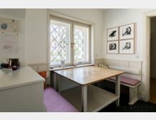 Kleine, gemütliche Essecke mit Holztisch und gepolsterten Sitzbänken unter einem Fenster mit verzierten Sprossen. An der Wand hängen vier gerahmte Vogelzeichnungen, daneben zieren Kinderzeichnungen die Wand.