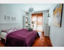 Schlafzimmer mit weißem Interieur, einem Doppelbett mit violetter Tagesdecke, Holzboden und Fenstern mit Holzjalousien. Im Raum stehen ein Heimtrainer und ein weißes Regal; an der Wand hängen Bilder und ein Blumenmotiv.