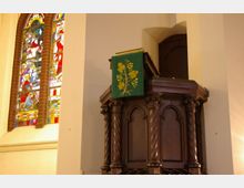 Holzkanzel mit geschnitzten Säulen und gotischen Bögen in einer Kirche, darauf ein grünes Tuch mit gelbem Traubenmotiv. Links ist ein buntes Glasfenster mit religiöser Darstellung zu sehen.