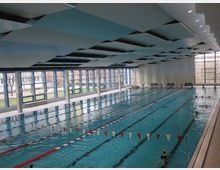 Innenraum eines modernen Schwimmbads mit einem großen Schwimmbecken, das durch Bahnleinen in mehrere Bahnen unterteilt ist. Große Fensterfronten lassen Tageslicht herein und bieten Blick auf eine Außenanlage mit Bäumen und Grünflächen.