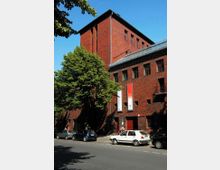 Backsteinbau im Stil der Moderne mit einem massiven Turm und rechteckigen Fenstern, umgeben von Bäumen. Vor dem Gebäude parken Autos entlang einer Straße bei sonnigem Wetter.