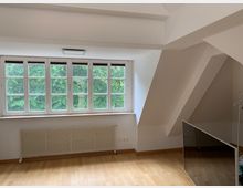 Dachzimmer mit weiß gestrichenen Wänden, einem großen Fenster mit Sprossen, das einen Blick auf grüne Bäume bietet, und einem Holzfußboden. Unter dem Fenster befindet sich ein Heizkörper, und rechts steht ein großer Spiegel, der an der Wand lehnt.