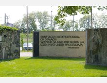 Denkmalanlage mit einer Wand, auf der ein Zitat von Karl Liebknecht in dunkler Schrift steht, umgeben von Mosaiken und Skulpturen. Im Hintergrund sind Bäume, Strommasten und ein Parkplatz zu erkennen, die Anlage liegt auf einer Grünfläche.