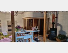 Gemütlicher Innenraum mit einer Sauna aus Holz an der Wand, einem Kaminofen mit Kupferrohr rechts und einer kleinen Bar mit dekorativen Vasen, Flaschen und Gläsern im Vordergrund. Links sind grüne Liegen und ein Weißholz-Regal zu sehen.