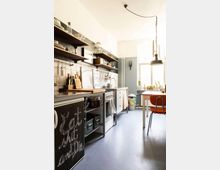 Helle Küche mit schlichten Möbeln und offenen Regalen, weißen Fliesen an der Wand und einem freistehenden Tisch mit orangefarbenem Stuhl. Im Raum sind Küchengeräte, Geschirr und Dekorationen sichtbar, während ein großes Fenster Tageslicht hereinlässt.