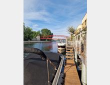 Ein Fluss mit einer roten Brücke im Hintergrund, flankiert von Bäumen auf der linken Seite und Gebäuden sowie einem Holzsteg mit festgemachten Booten auf der rechten Seite. Der Himmel ist klar mit einigen Wolken.