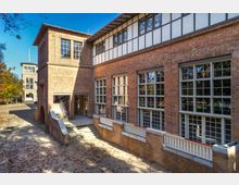 Backsteingebäude mit großen Fenstern im Stil der Moderne, umgeben von einem Hof mit Laub auf dem Boden. Seitlich führt eine Treppe mit Geländer hinauf, und im Hintergrund ist ein weiteres Gebäude mit ähnlicher Architektur zu sehen.