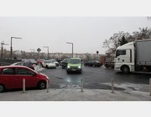 Kreuzung mit starkem Verkehr in einer städtischen Umgebung, umgeben von mehrstöckigen Wohngebäuden im Hintergrund. Verschiedene Fahrzeuge, darunter Pkw, ein Lieferwagen und ein Lkw, bewegen sich auf nasser Straße bei trübem Wetter.