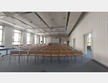 Großer Konferenzraum mit Reihen von beigen Stühlen auf einem blau-grauen Teppichboden, ein Projektorbildschirm und Lautsprecher an der Vorderwand; an den Seiten mehrere große Fenster und weiße, vertäfelte Wände.