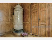 Eckbereich eines historischen Raumes mit Holzvertäfelung an den Wänden, einer geschlossenen Holztür rechts und einem verzierten, zylindrischen Kachelofen daneben. Auf dem Fischgrät-Holzfußboden steht ein kleiner, gepolsterter Fußschemel mit rosa Stoffbezug.