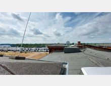 Blick von einem Flachdach auf eine ländliche Umgebung mit modernen weißen Wohnhäusern und einem Ziegelgebäude im Vordergrund. Im Hintergrund sind Grünflächen, eine Brücke und ein weitläufiger Himmel mit Wolken zu sehen.