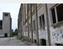 Verlassene Industriegebäude mit grauen Beton- und Ziegelwänden, teils mit Graffiti bedeckt; zahlreiche zerbrochene Fenster und verfallene Fassaden, umgeben von einem Betonhof mit vereinzeltem Pflanzenbewuchs.