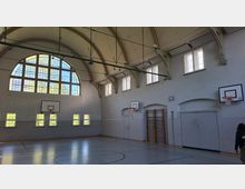 Innenansicht einer Sporthalle mit hohem Tonnengewölbe, großen Rundbogenfenstern und mehreren Basketballkörben an den Wänden. Der Hallenboden ist mit Spielfeldlinien markiert; an einer Seitenwand sind Sprossenwand und Türen zu sehen.