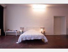 Ein schlicht eingerichtetes Schlafzimmer mit einem weißen Bett in der Mitte, flankiert von zwei Holzstühlen mit kleinen Tischlampen. Der Raum hat weiße Wände, einen dunklen Boden und ist spärlich dekoriert, mit einer Tür auf der rechten Seite.