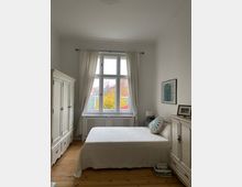 Ein helles Schlafzimmer mit einem großen Fenster in der Mitte, das von weißen Vorhängen eingerahmt ist. Zentral steht ein Bett mit weißer Tagesdecke, flankiert von einem weißen Schrank links und einer weißen Kommode rechts; der Holzboden ist sichtbar.