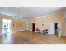 Ein heller, eleganter Saal mit Parkettboden, pastellfarbenen Wänden und einem Kronleuchter an der Decke. Große Fenster, weiße Türrahmen und ein Tisch mit Stühlen bestimmen das Interieur; der Raum wirkt aufgeräumt und ruhig.