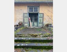 Eingang eines verwitterten Gebäudes mit einer alten, doppelflügeligen Holztür in Grün, umgeben von abblätternder Farbe. Eine kleine Treppe mit Moosbewuchs führt zum Eingang, und im Umfeld sind Pflanzen sowie der Eindruck eines vernachlässigten Gartens zu sehen.