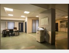 Bürobereich mit grünen Bodenfliesen und hellgrauen Wänden. Im Vordergrund steht ein Multifunktionsdrucker neben einer Wand mit einem Kalender, im Hintergrund sind ein Schreibtisch mit Bürostuhl sowie große Fenster mit Jalousien zu sehen.