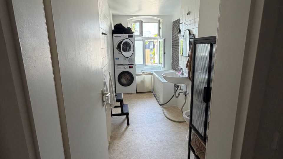 Schmaler Hauswirtschafts- und Waschraum mit weiß gefliesten Wänden, hellem Boden und einem Fenster am Ende. Links stehen übereinander Waschmaschine und Trockner, rechts sind Waschbecken, Badewanne und ein Duschbereich mit Glasabtrennung angeordnet.