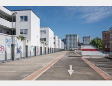 Moderne Wohnanlage mit weißen, geometrischen Gebäuden und flachen Dächern, flankiert von einem leeren Parkplatz mit markierten Stellflächen und Fahrbahn. Im Hintergrund sind mehrstöckige Büro- oder Wohngebäude sowie ein Treppenaufgang an einer Wand mit Graffiti zu sehen.