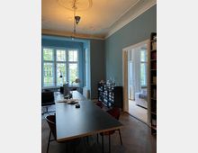 Ein stilvolles Büro in einem Altbau mit hohen Decken, verziertem Stuck und großen Fenstern, die viel Tageslicht hereinlassen. Der Raum verfügt über einen langen Tisch mit Stühlen, Regale mit Büchern und einen angrenzenden, lichtdurchfluteten Raum mit einem Sofa.