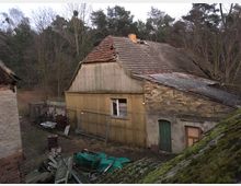 Verfallenes landwirtschaftliches Gebäude mit Ziegeldach, teilweise beschädigt, und Wänden aus gelben und braunen Ziegeln. Im Vordergrund befinden sich Schutt, alte Gegenstände und eine Badewanne; im Hintergrund steht ein dichter Wald.