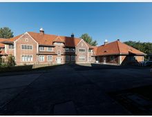 Großer Backsteinbau mit roten Ziegeldächern und mehreren kleinen Dachaufsätzen, frontseitig mit vielen rechteckigen Fenstern und einem mittigen Eingangsbereich. Davor liegt ein gepflasterter Hof, umgeben von Bäumen unter klarem blauem Himmel.