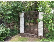 Verrostetes schmiedeeisernes Gartentor mit ornamentalen Mustern zwischen zwei hellen Steinpfeilern, umgeben von dichtem Grün und Bäumen. Dahinter ist ein schmaler Gartenweg und eine niedrige Betonmauer mit dekorativen Aussparungen zu sehen.