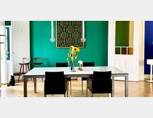 Essbereich mit einem weißen Tisch, vier schwarzen Stühlen und einer Vase mit Sonnenblumen. Der Raum hat eine grüne Wand mit einer dekorativen Holzstruktur, hängenden Lampen, sowie eine Tür mit bunt verglasten Fenstern.