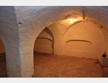 Ein kleiner, gewölbter Kellerraum mit weiß verputzten Wänden und einem einfachen Betonboden. Die Decke ist durch Rundbögen gestützt, und eine einzelne, hängende Glühbirne beleuchtet den Raum.