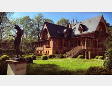 Historische Villa aus rotem Backstein mit Fachwerkelementen und einem Schieferdach, umgeben von einem gepflegten Garten mit Rasenflächen, Sträuchern und einer Statue im Vordergrund. Im Hintergrund sind hohe Bäume sichtbar.