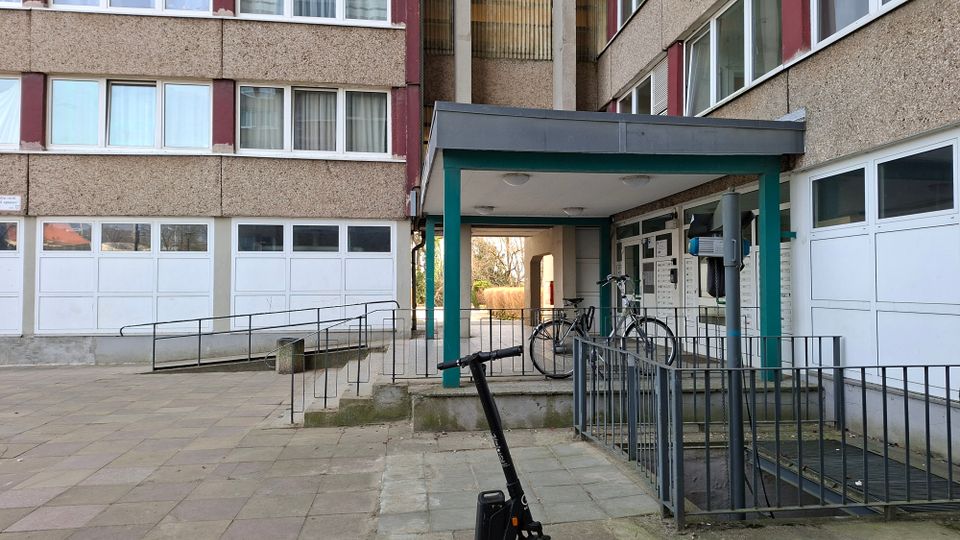 Eingangsbereich eines mehrstöckigen Wohnblocks mit rauer Fassadenstruktur, großen Fensterreihen und einer überdachten Vorfahrt mit grünen Stützen. Davor liegen gepflasterte Wege mit Rampe und Geländern sowie ein Fahrradabstellbereich neben den Haustürbriefkästen.