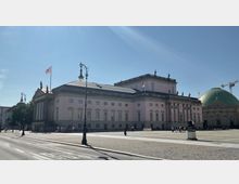 Historisches Gebäude mit klassizistischer Architektur, gekennzeichnet durch Säulen und Statuen auf dem Dach, neben einer großen Kirche mit grüner Kuppel auf einem offenen Platz. Der Himmel ist klar, und die Straße im Vordergrund ist wenig befahren.