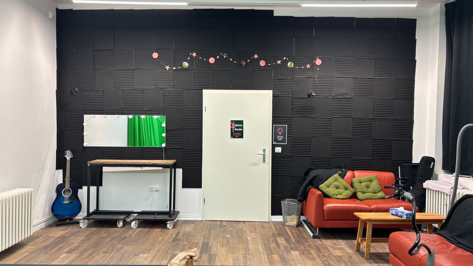 Interior of a small studio lounge with a black acoustic foam wall and a central white door, wood flooring, and bright fluorescent ceiling lights. A red leather sofa with cushions sits beside a small wooden coffee table, with a vanity-style mirror and table on wheels and a guitar along the left wall.