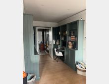 Ein Arbeitszimmer mit grün gestrichenen, eingebauten Regalen und Schränken an den Wänden. In der Mitte steht ein weißer Stuhl an einem Schreibtisch; der Raum hat helle Holzböden und eine Tür führt in einen angrenzenden Flur.