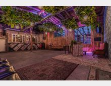 Industrie-inspirierter Innenraum einer Bar oder eines Veranstaltungsortes mit Holzwänden, freiliegenden Deckenbalken und hängenden Pflanzen. Im Raum befinden sich gemütliche Sitzbereiche, Teppiche, eine Theke mit Barhockern sowie große Fenster mit Blick nach draußen.