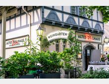 Fassade eines historischen Fachwerkhauses mit den Schildern „Weingarten“ und „Altes Zollhaus“, mit Laternen an der Wand und einem Rundbogen-Eingang. Davor stehen üppige Grünpflanzen in Kübeln und ein kleiner Außenbereich mit Tischen im Hof.