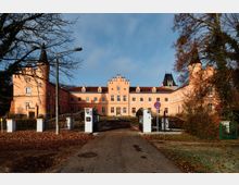 Frontansicht eines rosafarbenen Schloss- oder Herrenhauskomplexes mit zentralem Giebel und seitlichen Türmen, erreichbar über eine Zufahrt durch ein schmiedeeisernes Tor. Vor dem Gebäude stehen weiße Torpfeiler und Laternen, umgeben von Bäumen und herbstlichem Laub.